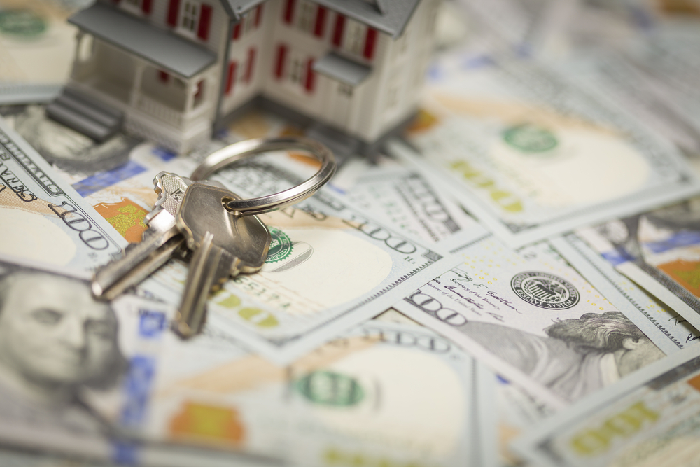 A small model house and set of keys positioned on several crisp, newly designed U.S. one hundred dollar bills, symbolizing the concept of 'cash for homes' or selling a home for immediate cash.