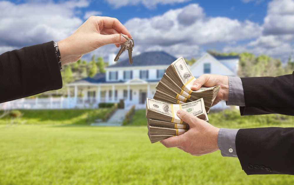 Hands exchanging house keys for a cash offer with a beautiful home in the background, symbolizing a sell home cash transaction.